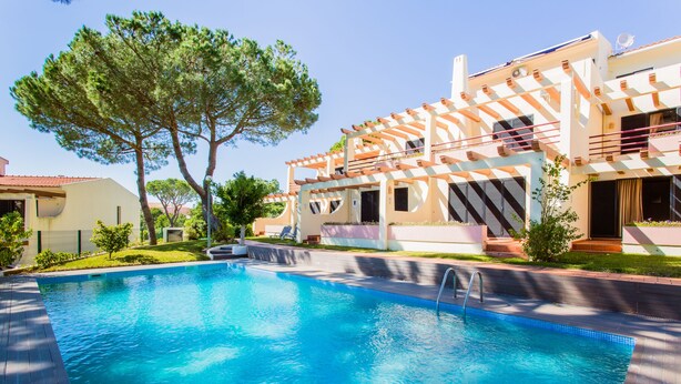 A swimming pool with a wooden deck, surrounded by a two-story building with balconies and a large tree. - Beach House (Golf) - Falésia/Albufeira (Olhos Dagua)