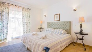 A neatly arranged bedroom with a large bed, two bedside tables with lamps, and a window with floral curtains. - Beach House (Golf) - Falésia/Albufeira (Olhos Dagua)