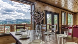 Taula de menjador preparada amb plats, gots i un ram de flors, amb vista a les muntanyes a través de grans finestres.