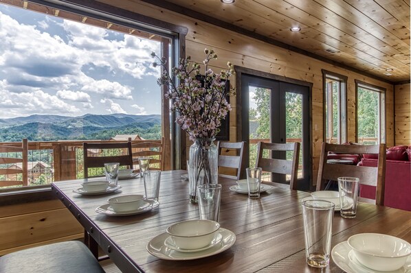 A dining table set with plates, glasses, and a vase of flowers, with a view of mountains through large windows.