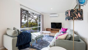 A modern living room with a large window, a flat-screen TV, and a decorative lamp. - WindanSea Cottage - Pet Friendly - Ocean Glimpses (Vincentia)