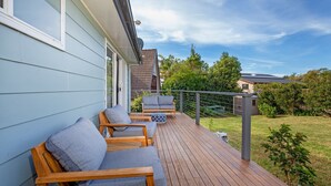 A wooden deck with a sofa and chairs, a metal railing, and a view of a garden and neighboring houses. - WindanSea Cottage - Pet Friendly - Ocean Glimpses (Vincentia)