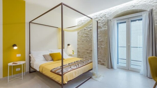 Ein Himmelbett mit gelber Bettdecke und Kissen, ein kleiner Nachttisch mit Lampe und Blick ins Freie durch eine Glastür.