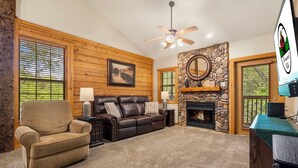 A cozy living room with a fireplace, a stone wall, and a ceiling fan. - Peaceful Walk-in Cabin with Whirlpool Tub, Gas Fireplace, and Screened-in Porch (Branson)