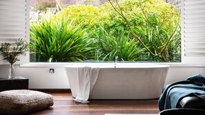 A modern bathroom with a freestanding bathtub, a window with a view of lush greenery, a small bedside table, and a cozy seating area. - Chestnut Daylesford-Log fire place, spa bath and close to all of Daylesford's attractions (Daylesford)
