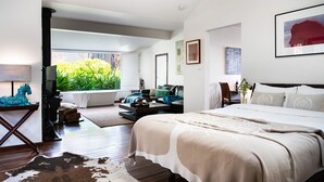 A modern bedroom with a large bed, a sitting area with a sofa and coffee table, a window with a view of greenery, and a bathroom with a bathtub. - Chestnut Daylesford-Log fire place, spa bath and close to all of Daylesford's attractions (Daylesford)