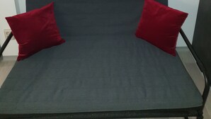 A grey sofa with two red cushions against a white wall, a black-framed wall clock above it, and a black metal frame on the right. - Relaxhaus Kehl (Kehl)