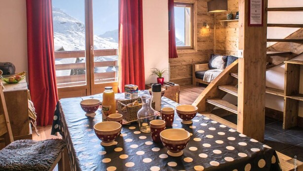 Une pièce chaleureuse avec un escalier en bois, une table à manger dressée avec des bols et une bouteille, et une vue sur des montagnes enneigées par de grandes fenêtres.