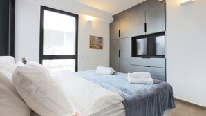 A modern hotel room with a large bed, a flat-screen TV mounted on the wall, and a window providing a view of the city. - AirTLV - Nes Tziona Luxury By The Sea (Tel Aviv)