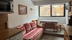 A compact living space with a kitchenette, a sofa with patterned cushions, a dining table, and a large window offering a view of buildings. - 50m from resort center, 6th floor, tv, 27m², Val Thorens (Val Thorens)