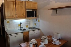 Cocina compacta con muebles de madera, microondas, lavamanos y un pequeño comedor con tazas y hervidor.
