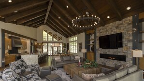 A spacious living area with a fireplace, a large TV mounted on a stone wall, and a modern kitchen in the background. - One-of-a-Kind Luxury Home - Hot Tub, Firepit, Views - 8 Min to Purg (Durango)