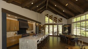 A modern kitchen with a large island, wooden cabinets, and a view of the outdoors through large windows. - One-of-a-Kind Luxury Home - Hot Tub, Firepit, Views - 8 Min to Purg (Durango)