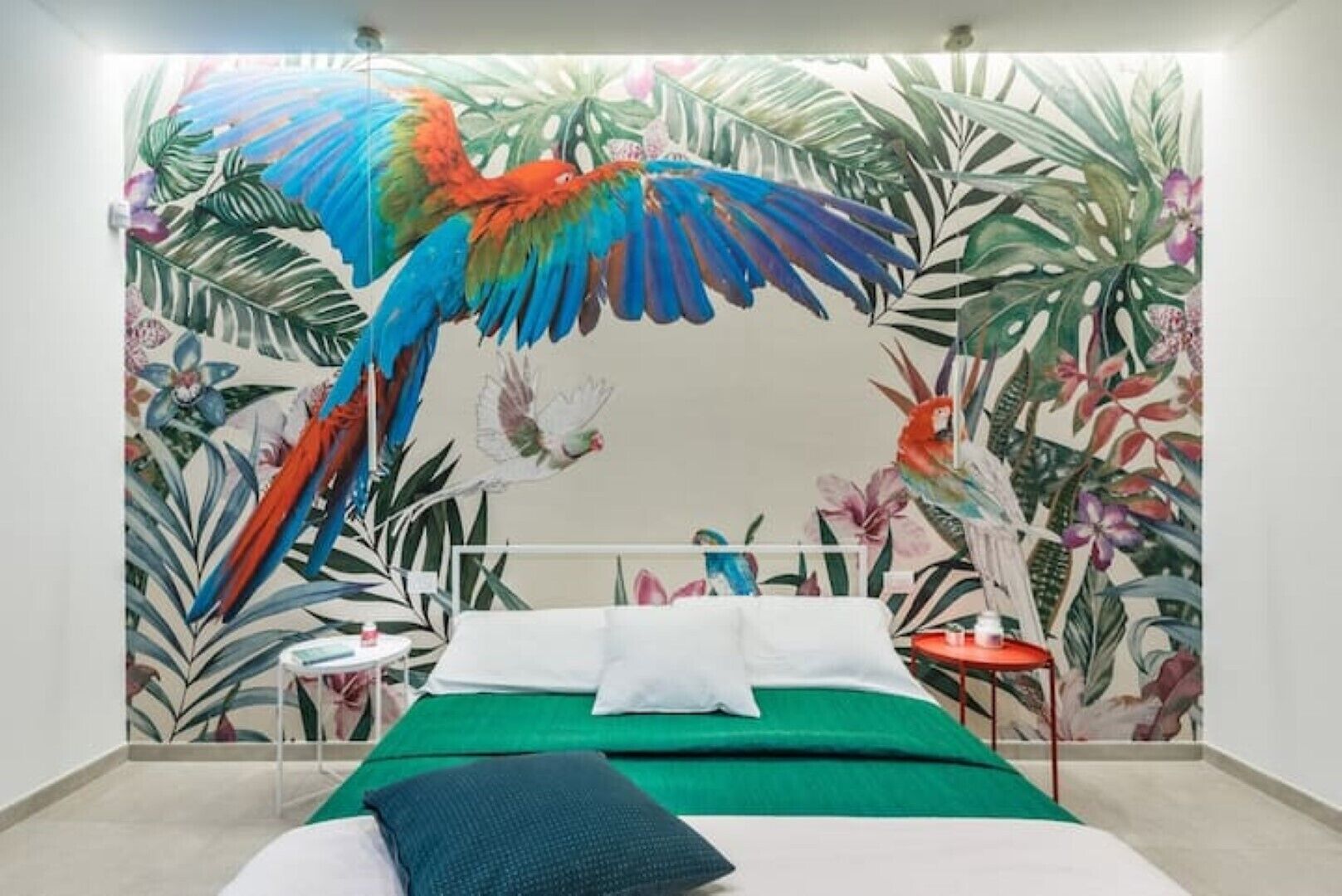 A bedroom with a bed, green bedding, a white pillow, a red pillow, a nightstand with a lamp, and a wall mural of tropical leaves and birds.