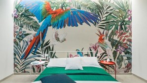 Une chambre à coucher avec un lit, une literie verte, un oreiller blanc, un oreiller rouge, une table de chevet avec une lampe, et une fresque murale représentant des feuilles et des oiseaux tropicaux.