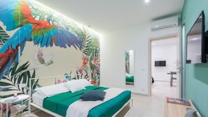 Une chambre moderne avec un grand lit, une table de chevet, un miroir et un mur orné d’une fresque aux motifs tropicaux.