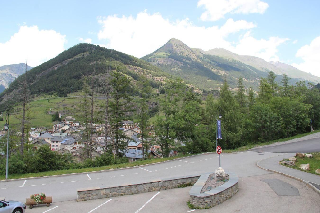A mountain road with a village in the valley, a no parking sign, and a stone monument.