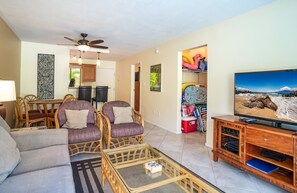 A living room with a sofa, two armchairs, a coffee table, a TV on a wooden stand, and a ceiling fan.
