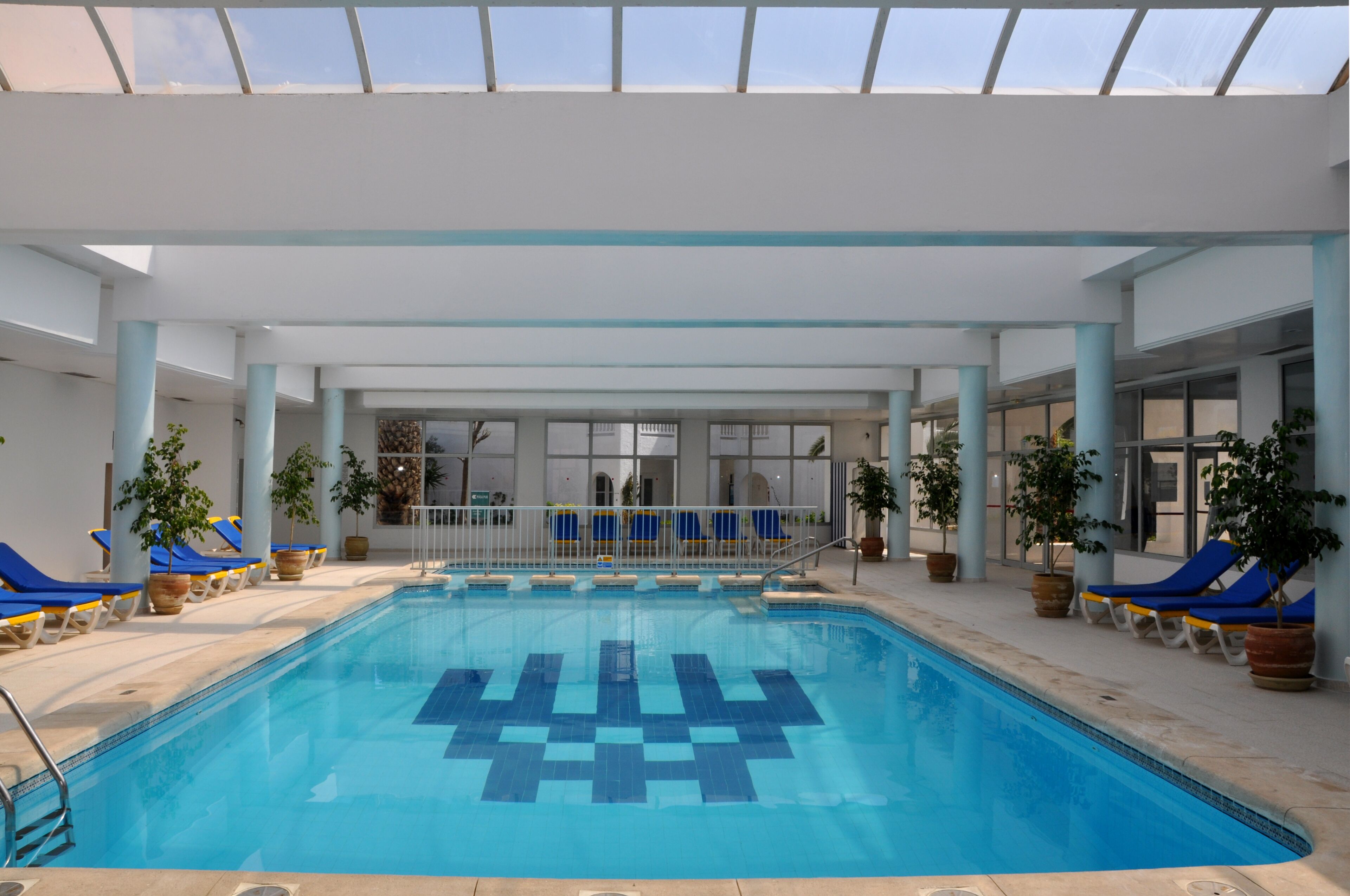 Una piscina interior con sillones reclinables azules y amarillos, plantas en macetas y techo de cristal.