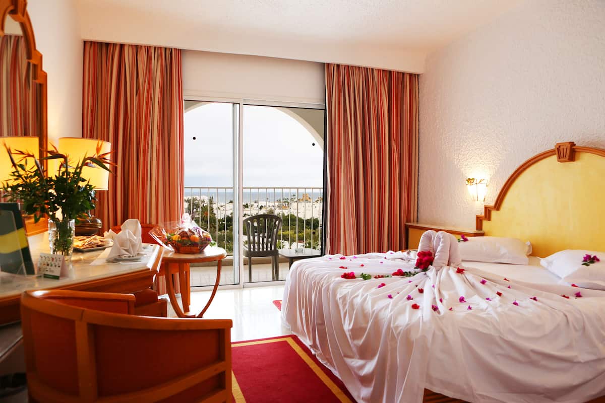 Een hotelkamer met een bed, een bureau, een stoel, een tafel en een uitzicht op de stad door grote ramen.