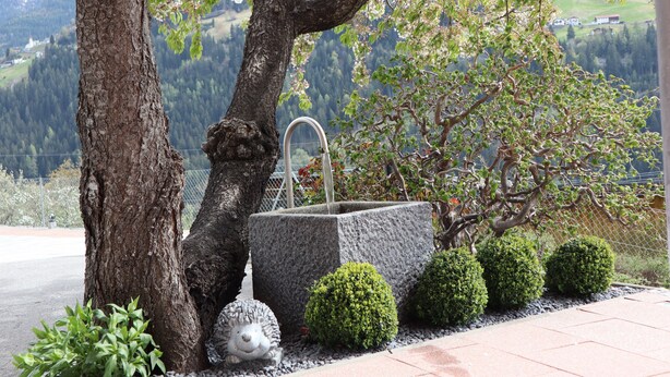 Una fontana in pietra con un beccuccio metallico, circondata da siepi curate e una statuina di riccio.
