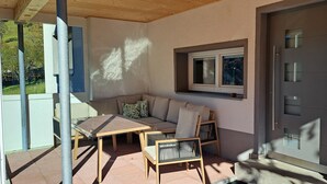 Un’area salotto esterna coperta, con tavolo e sedie, un divano e una finestra con persiane.