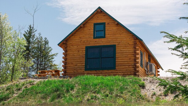 A log cabin with a large window and a door, surrounded by a grassy area and trees.