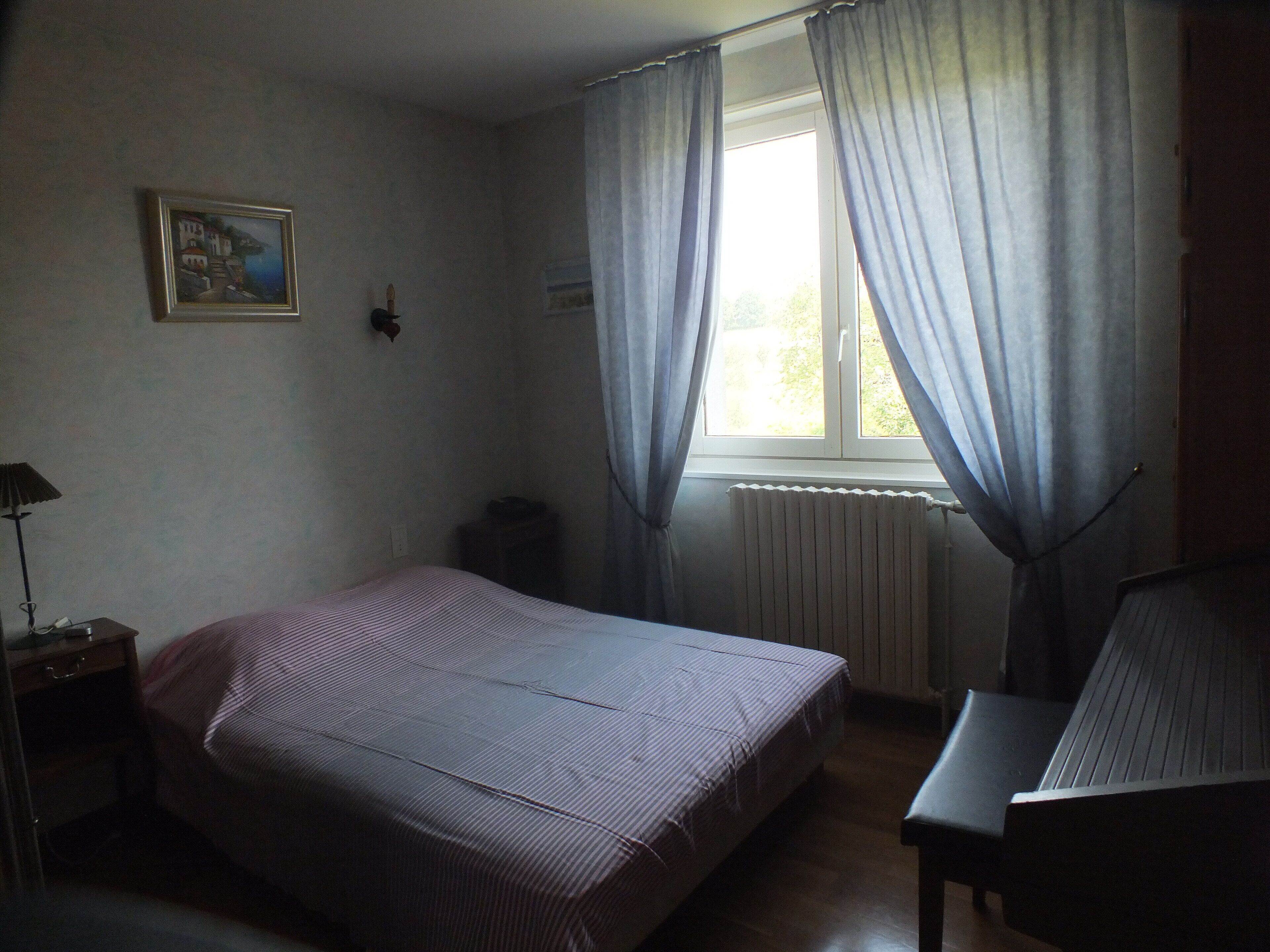 Une chambre à coucher comprenant un lit, une chaise, une table de chevet, une lampe, un radiateur et une fenêtre avec des rideaux.