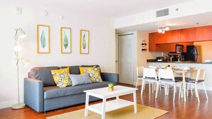 A modern living room with a grey sofa, white coffee table, and a dining area with white chairs and a kitchen in the background. - 2BR + Rooftop Pool | Free Parking Included (Miami)