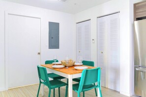 Ein Essbereich mit einem Holztisch, grünen Stühlen und einem Brotkorb. Weiße Türen und ein kleines Fenster mit blauem Rollo sind zu sehen.