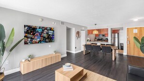 Moderni olohuone, jossa on suuri televisio, puuhuonekaluja ja ruokailutila, jonka taustalla näkyy keittiö.