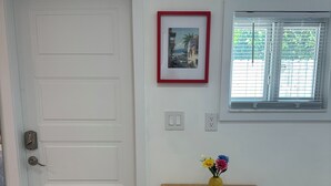 Une pièce avec une porte blanche, un tableau encadré au mur, une fenêtre avec des stores, une table avec un set de table, un vase avec des fleurs et un paillasson devant la porte.