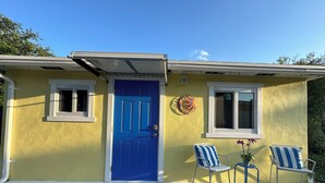 Un bâtiment jaune avec une porte bleue, deux fenêtres et un petit porche agrémenté de deux chaises et d’une plante en pot.