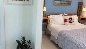 Une chambre à coucher avec un lit, une table de chevet, une plante en pot, un tableau accroché au mur et une étagère avec un vase et des fleurs.