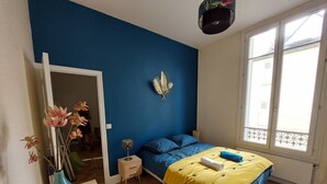 A bedroom with a blue wall, a bed with a yellow blanket, a nightstand, a vase with flowers, and a window with a view.