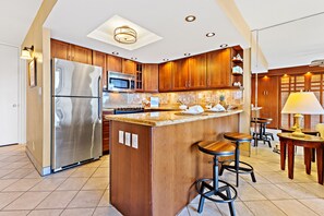 A modern kitchen with wooden cabinets, stainless steel appliances, a central island with bar stools, and a dining area with a table and chairs. - Hawaiian-inspired studio with ocean view (Lahaina)