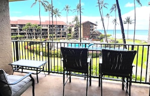 Un balcon avec une table et deux chaises, donnant sur une piscine et une plage bordée de palmiers.