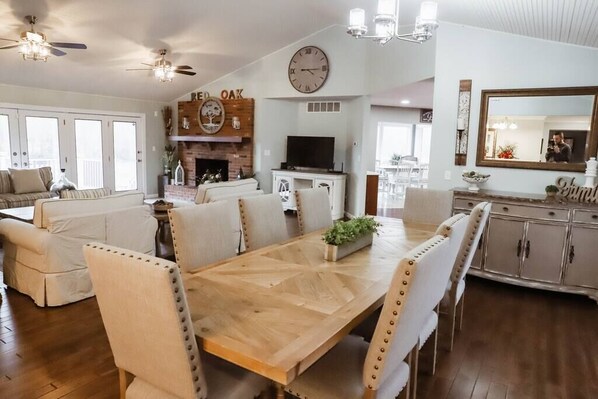 A spacious living room with a wooden dining table, comfortable seating, a fireplace, and a large clock on the wall.