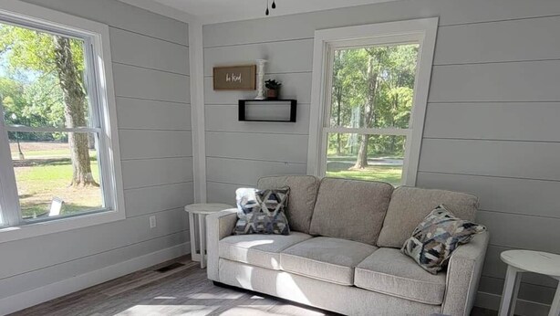 A spacious living room with a grey sofa, a grey armchair, and a small white table. There is a ceiling fan, a window with a view of trees, and a shelf with a sign saying "Be kind." - #3 Cabin at Red Oak Valley (Canaan Township)