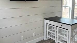 Una habitación con paredes blancas empañadas, un televisor de pantalla plana montado, una mesa blanca con cajones y una ventana con vistas a árboles.