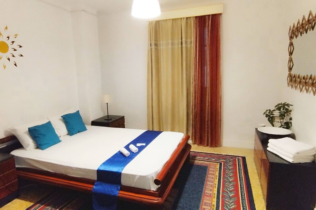 A hotel room with a bed, a nightstand, a lamp, a mirror, a curtain, and a small plant.
