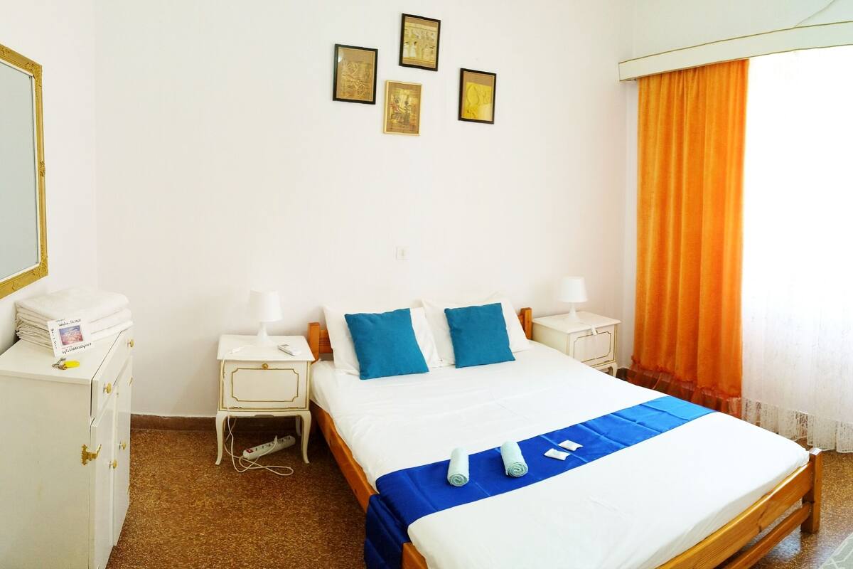 A neatly made bed with blue and white bedding, a bedside table with a lamp, and a window with orange curtains.