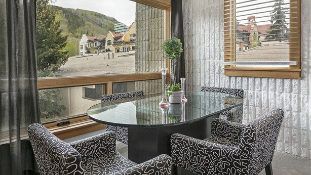 A dining area with a round glass table and patterned chairs, a potted plant, and a view of a town and mountains through large windows.