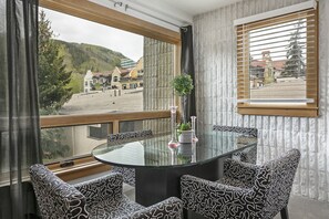 Un comedor con una mesa redonda de vidrio y sillas estampadas, una planta en maceta y vistas a un pueblo y montañas a través de amplios ventanales.