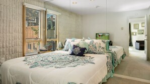 A bedroom with a large bed, a window with blinds, and a door leading to another room.