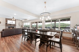 Un comedor amplio con una mesa larga y numerosas sillas, una chimenea y ventanas grandes que ofrecen vistas al exterior.