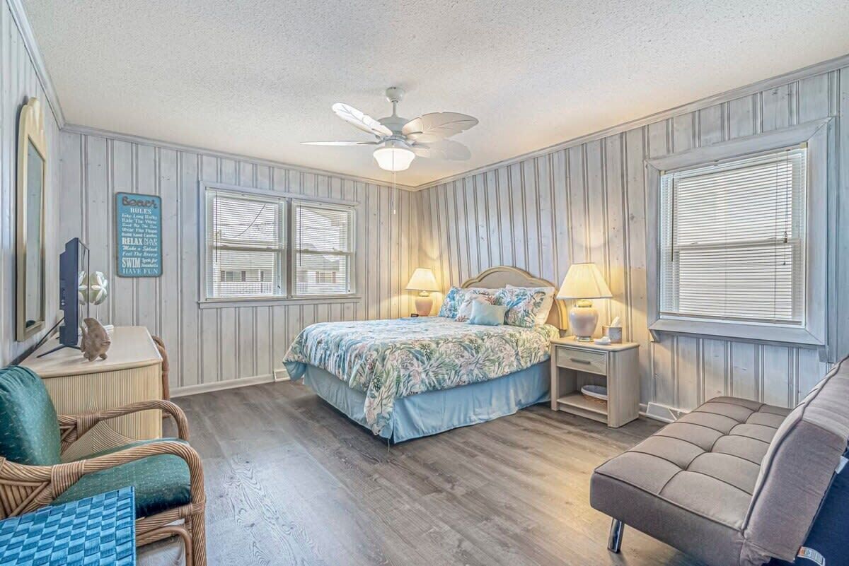 Een slaapkamer met een bed, twee nachtkastjes, een stoel, een bank, een plafondventilator en een raam met jaloezieën.