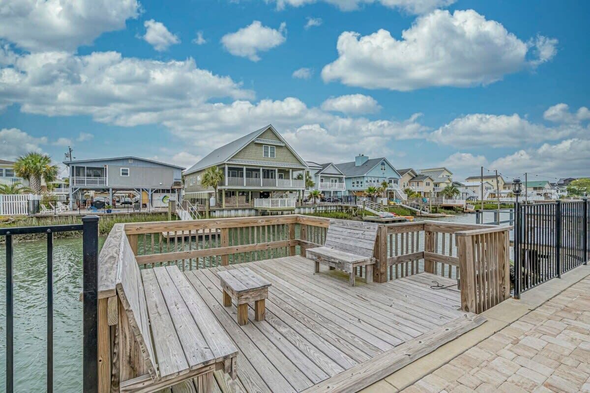 A wooden dock with benches, a small table, and a view of houses along a waterfront.