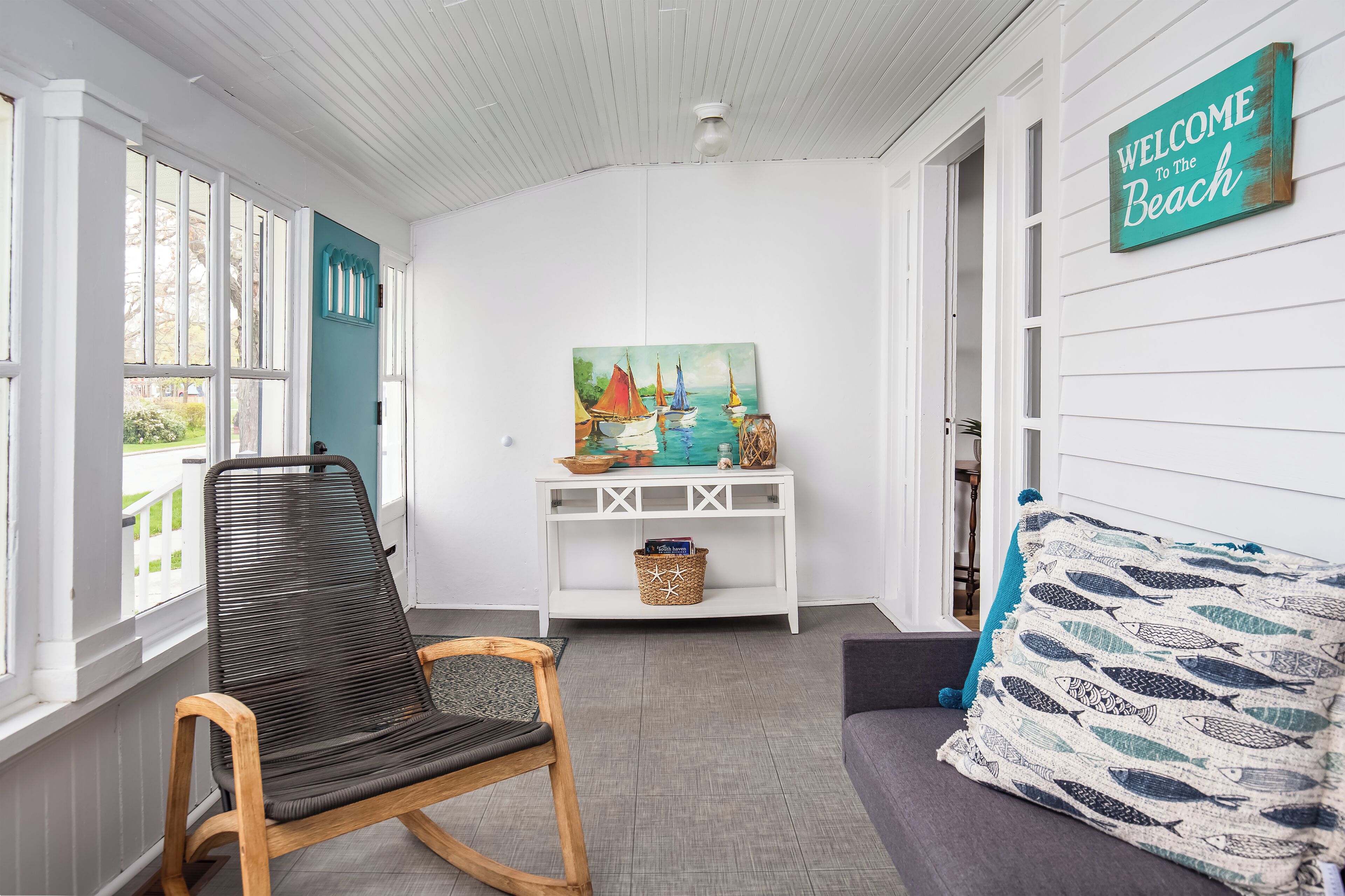 Une pièce avec un fauteuil à bascule, une console sur laquelle est posée une peinture, un panneau indiquant « Bienvenue à la plage », et un canapé gris agrémenté d’un coussin à motifs.