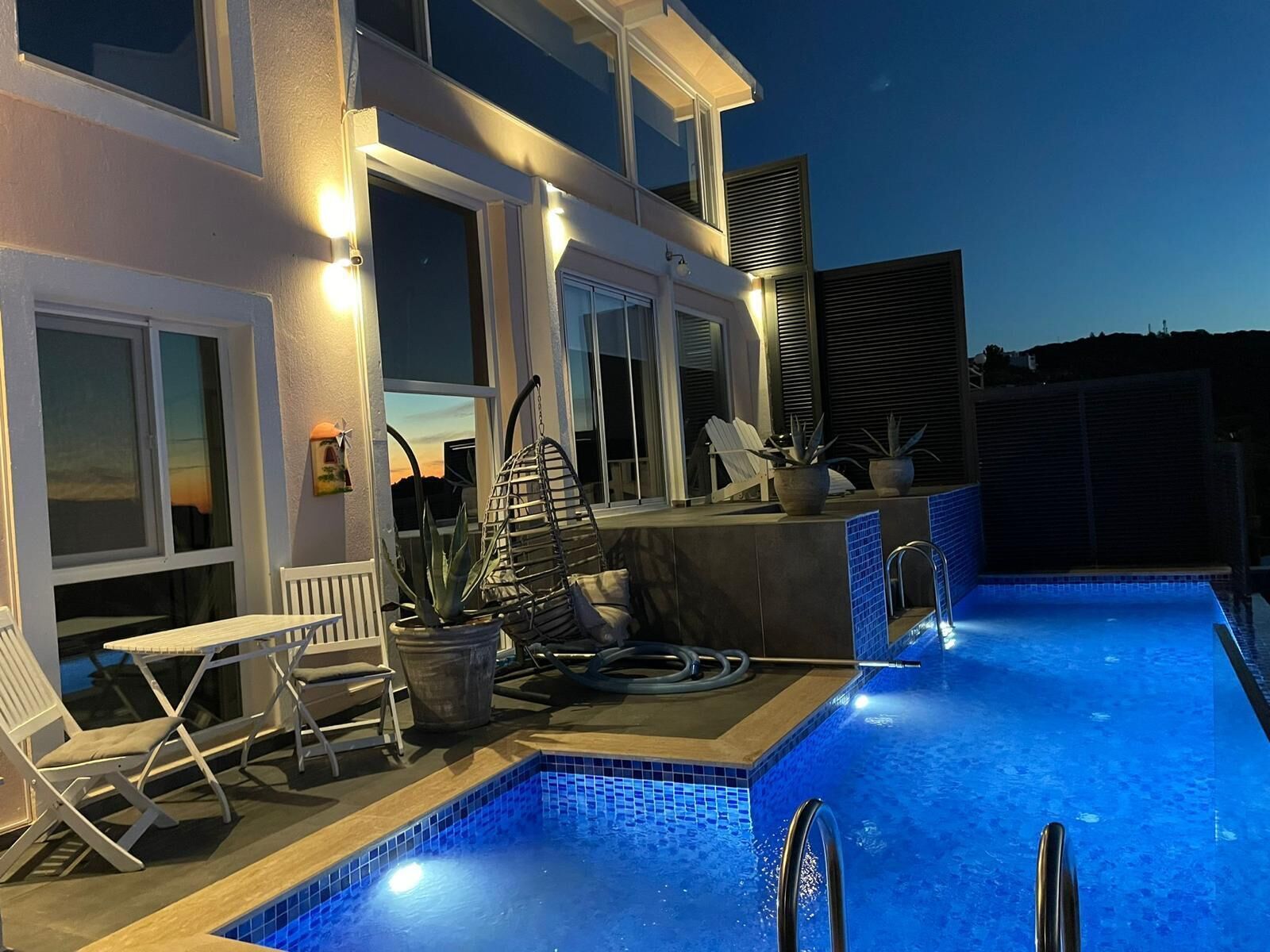 Une piscine avec un bord carrelé, un escalier en métal et un espace extérieur aménagé avec des chaises et une table, le tout de couleur blanche.